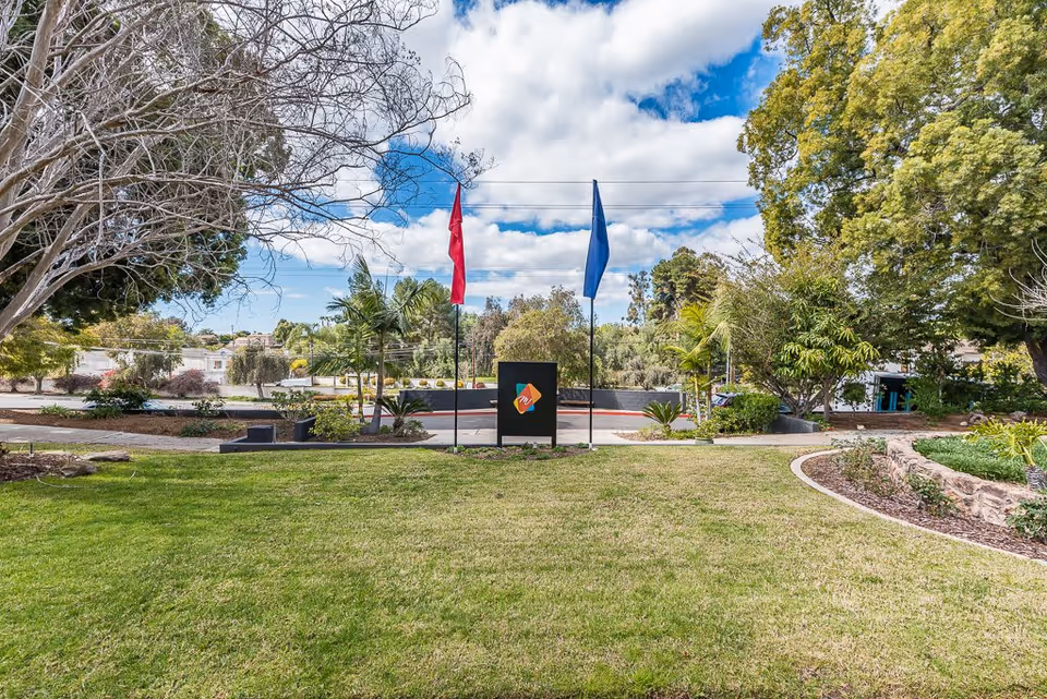 A landscaped outdoor area with green grass, trees, and shrubs under a partly cloudy blue sky. Two flagpoles with red and blue flags flank a black sign with a colorful logo in the center. Surrounding the area are various plants and a stone-bordered garden bed.
