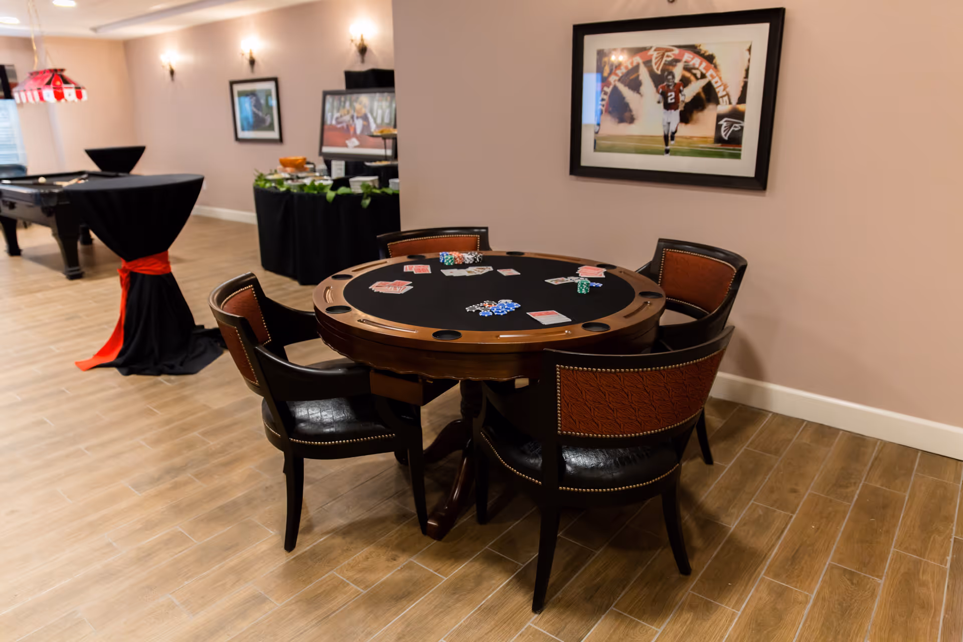 A game room with a round poker table surrounded by five chairs, poker chips and cards on the table. In the background, there is a pool table, a high round table covered with a black cloth and a red sash, and a buffet table with food. The walls are decorated with framed sports pictures.