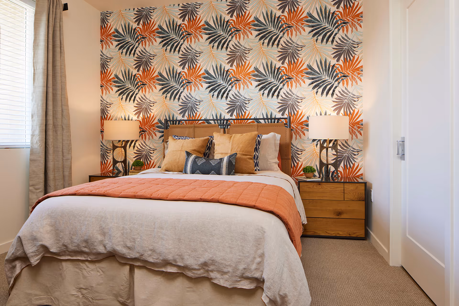 A neatly made bed with decorative pillows and an orange throw against a bold tropical-patterned accent wall, flanked by wooden nightstands and lamps.