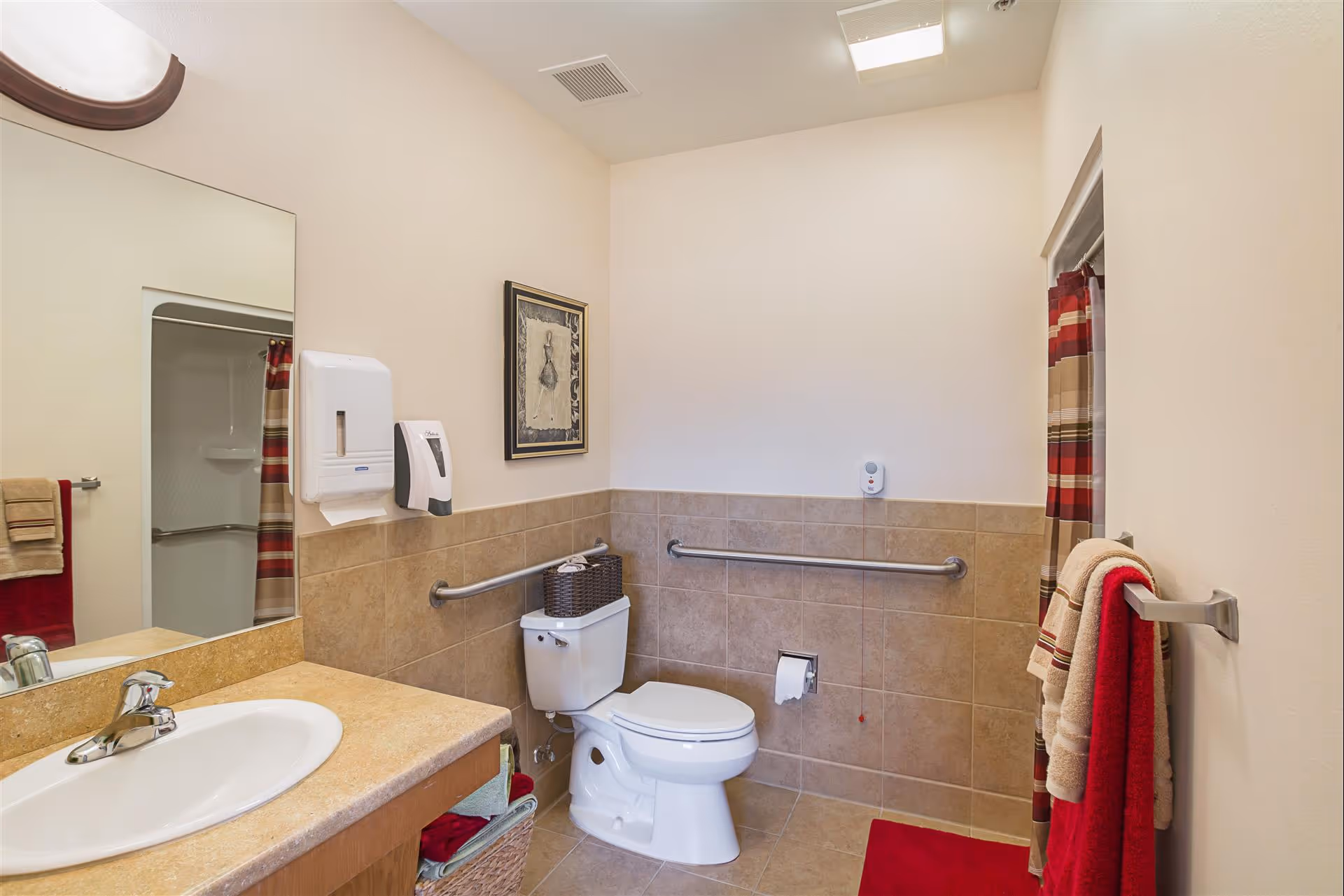 A clean and accessible bathroom with beige tiled walls and floor. It features a white toilet with grab bars on both sides, a beige countertop with a white sink, a large mirror above the sink, a wall-mounted soap dispenser and paper towel holder, a framed picture on the wall, a red and beige striped shower curtain, and red and beige towels hanging on a towel rack.