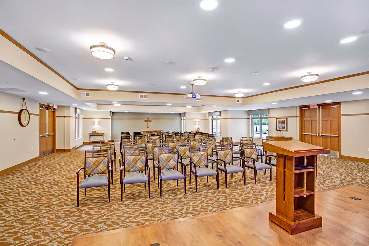 A spacious meeting or chapel room with rows of wooden chairs with patterned cushions facing a wooden podium. The room has beige walls, patterned carpet, multiple ceiling lights, a wall clock, and a cross mounted on the far wall. There are double doors and windows letting in natural light.