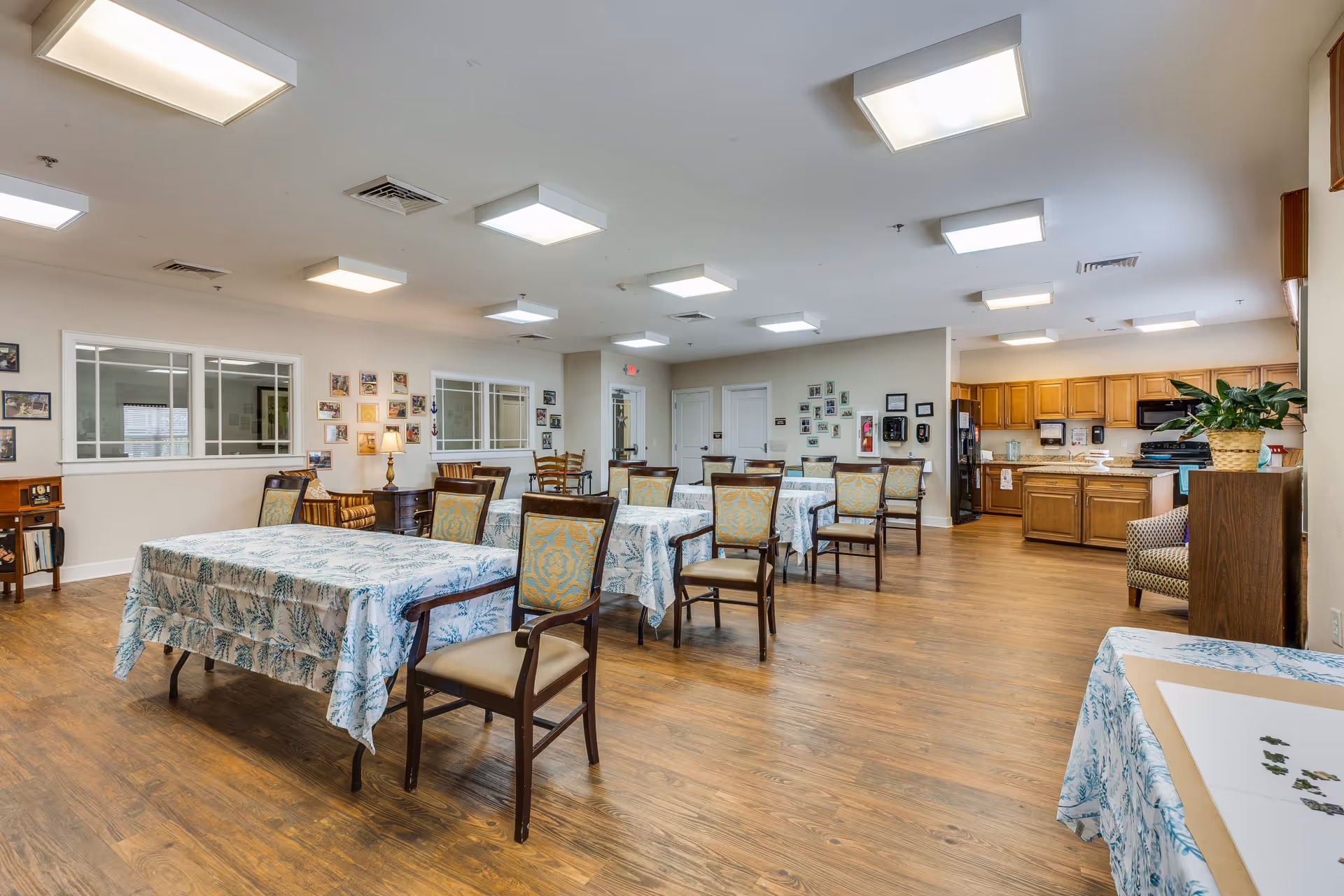 A spacious dining area in an assisted living facility with several tables covered in patterned tablecloths and surrounded by wooden chairs with cushioned seats. The room has wooden flooring, bright ceiling lights, and walls decorated with numerous framed photos. In the background, there is a kitchen area with wooden cabinets, a refrigerator, and a stove. A potted plant sits on a piece of furniture near the kitchen.