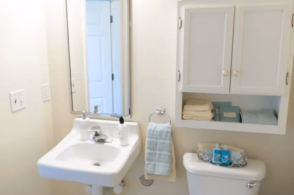Small bathroom with a pedestal sink, mirror, wall cabinet, towel and a toilet topped with toiletries.
