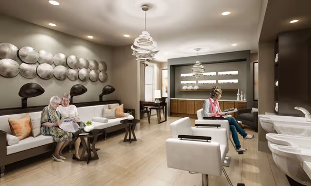 A modern senior living lounge area with two elderly women sitting on a white sofa reading a newspaper, and another woman sitting in a salon chair reading a magazine. The room features stylish decor including circular wall art, pendant lights, and a counter with shelves displaying decorative items.