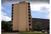 Multi-story beige senior living building with a central tower and adjoining wings under a partly cloudy sky.