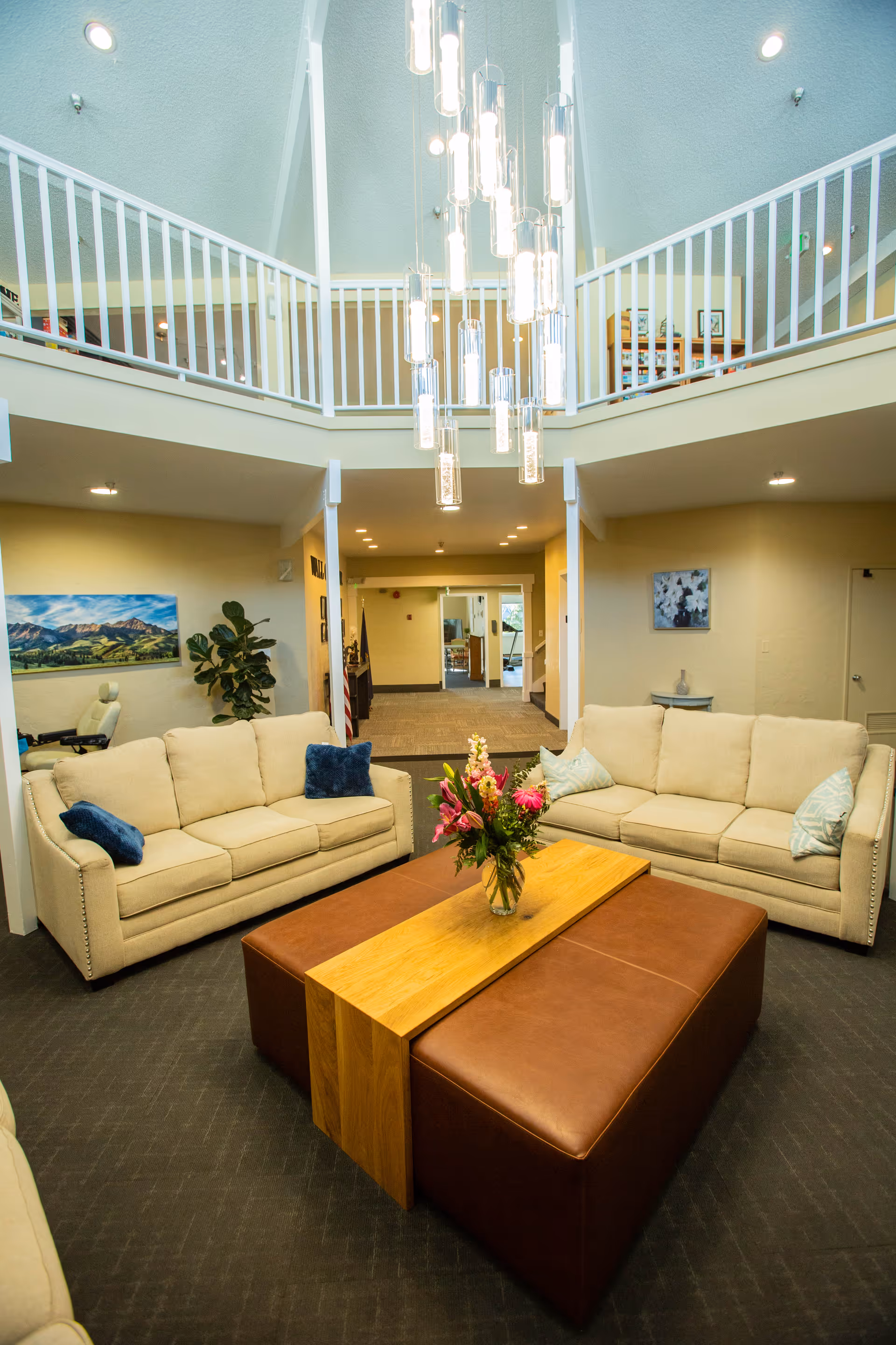 A bright and spacious senior living facility common area with two beige sofas adorned with blue and light-colored pillows facing a large rectangular coffee table with a wooden centerpiece and brown leather sides. A vase with colorful flowers sits on the table. The room has a high ceiling with a modern chandelier hanging down. There is a second-floor balcony with white railings overlooking the area. The walls are light-colored, and there are paintings and plants decorating the space.