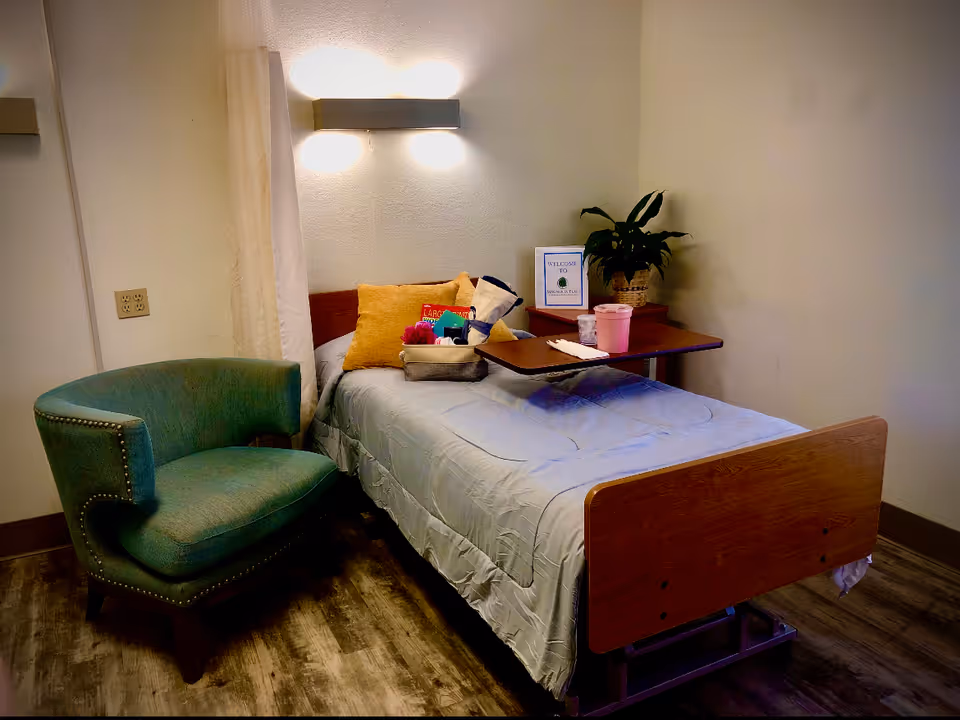 A small patient bedroom with a single bed, overbed table, green upholstered chair, wall light, and a bedside plant and welcome sign.