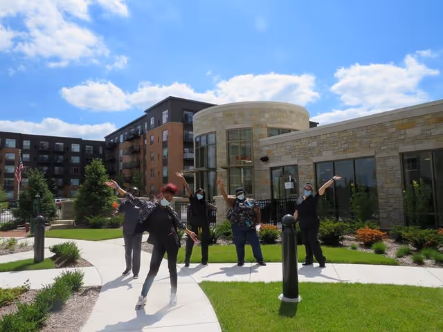 Five people wearing masks standing outside on a paved walkway in front of a modern building with stone and brick exterior, waving and posing under a partly cloudy blue sky.