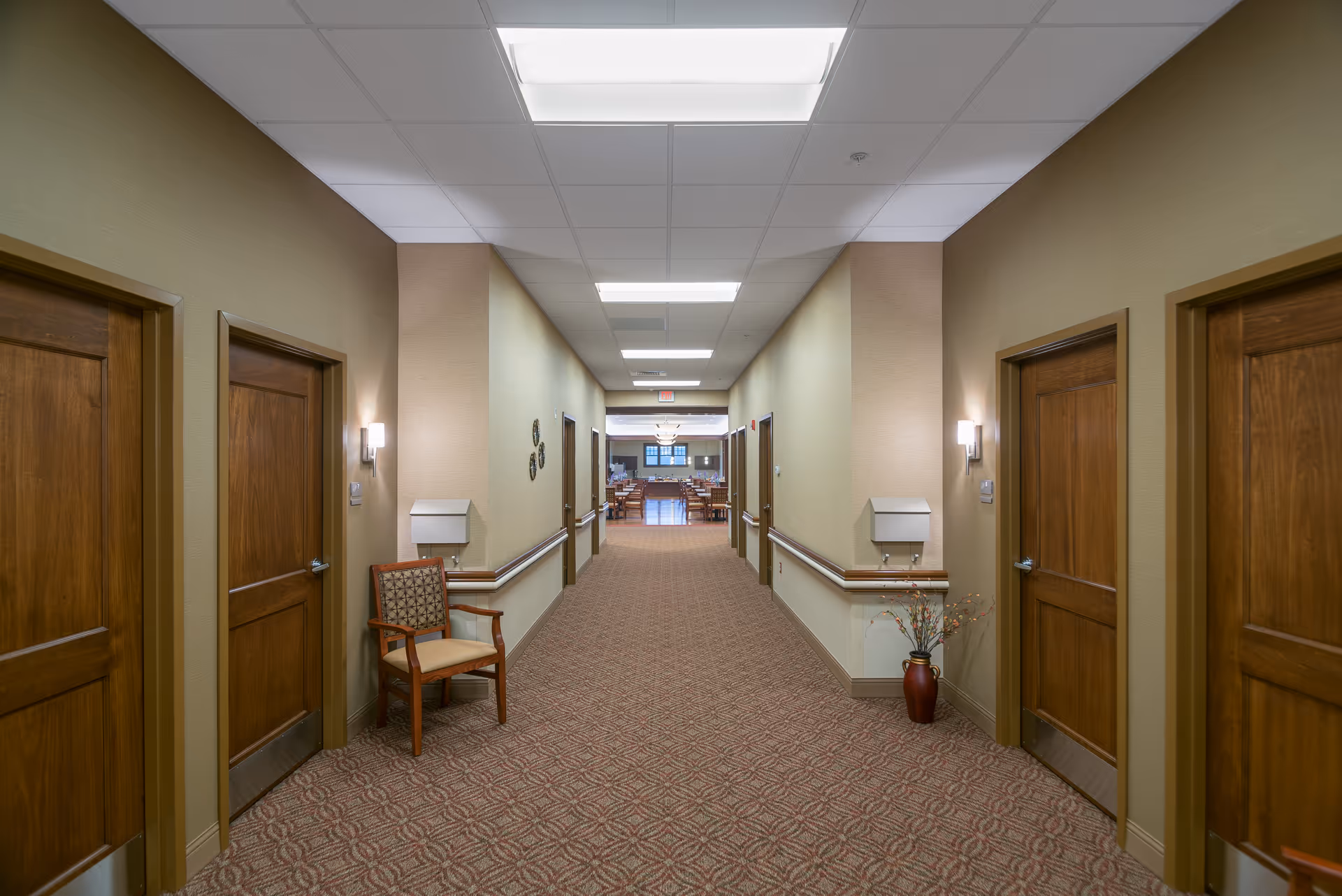 A well-lit hallway in an assisted living facility with beige walls, patterned carpet, wooden doors on both sides, a wooden chair with a patterned cushion on the left, and a vase with decorative branches on the right. At the end of the hallway, a dining area with tables and chairs is visible.