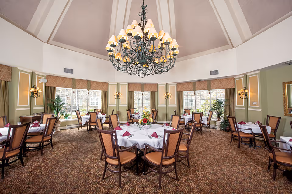 A spacious dining room with round tables covered in white tablecloths and set with burgundy napkins, glassware, and floral centerpieces. The room features large windows with beige curtains, green walls with decorative molding, and a large ornate chandelier hanging from a high ceiling.
