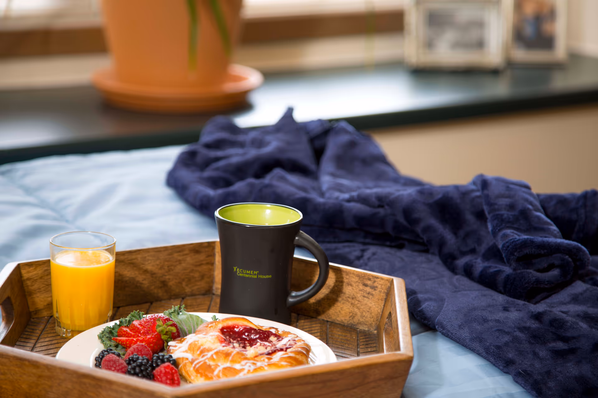 A wooden tray on a bed holding a plate with a pastry topped with icing and jam, fresh berries, a glass of orange juice, and a black mug with a green interior labeled 'Ecumen Centennial House'. A dark blue blanket is draped on the bed near the tray, with a window and a potted plant in the background.