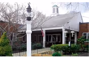 Exterior view of a nursing home building with a covered entrance, an American flag, a white lamp post, and surrounding greenery including bushes and trees.