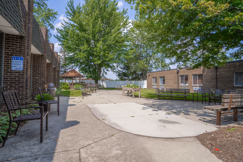 Outdoor courtyard area at Mission Point of Clinton Township with benches, chairs, potted plants, a gazebo, and large trees providing shade. The area is surrounded by brick buildings and has a designated smoking area sign on one wall.