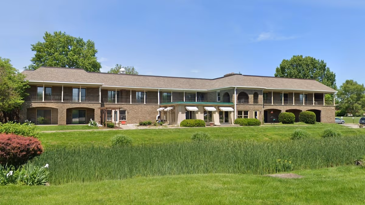Two-story brick assisted living building with covered balconies facing a grassy lawn and small pond under a clear blue sky.