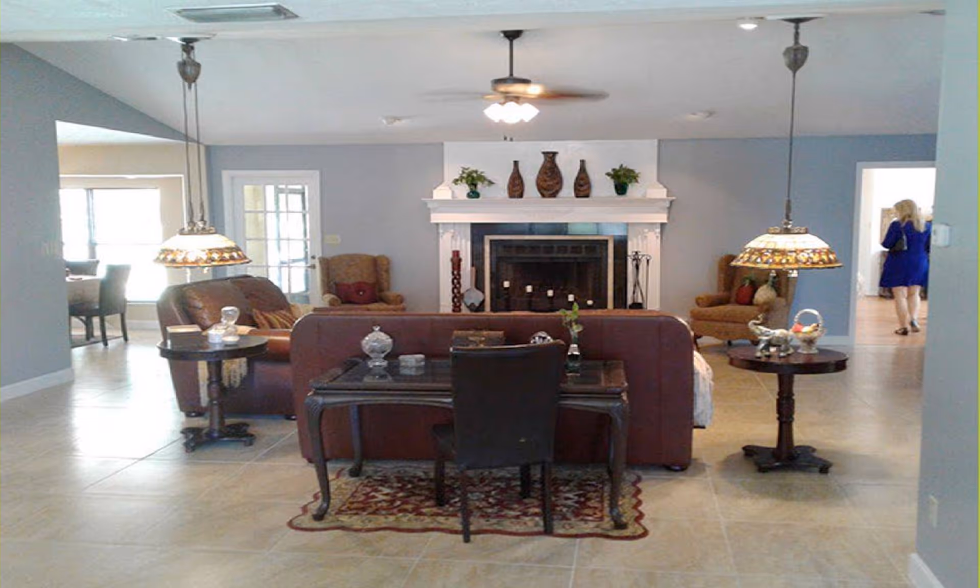 A spacious living room with a brown leather sofa facing a white fireplace adorned with decorative vases and plants. Two armchairs are positioned on either side of the fireplace. There are two round side tables with lamps hanging above them. A desk with a chair is placed behind the sofa on a patterned rug. The room has tiled flooring and light blue walls. In the background, a woman in a blue dress is walking through a doorway.