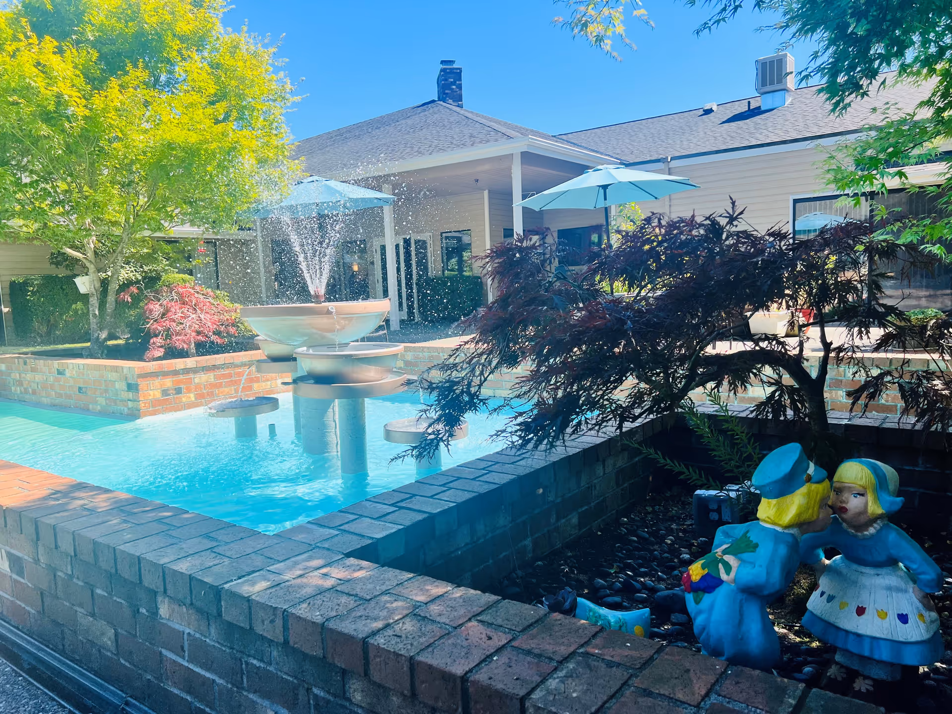 Outdoor courtyard area at Cap Sante Court Retirement Community featuring a brick-bordered water fountain with water spraying upwards, surrounded by trees and plants. There are blue umbrellas providing shade over seating areas, and a decorative statue of two children, one dressed as a mail carrier and the other as a girl, near the fountain.