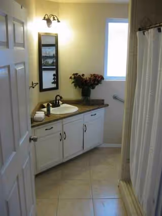Bathroom with white vanity, sink, mirror, flowers, and a shower in a senior living facility.