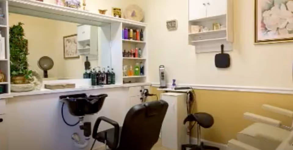 Interior view of a small salon area in an assisted living facility with a black salon chair in front of a black wash basin, shelves with various hair care products, a large mirror, and additional seating including a small black stool and beige chairs. The walls are painted white and yellow with a framed floral picture hanging.