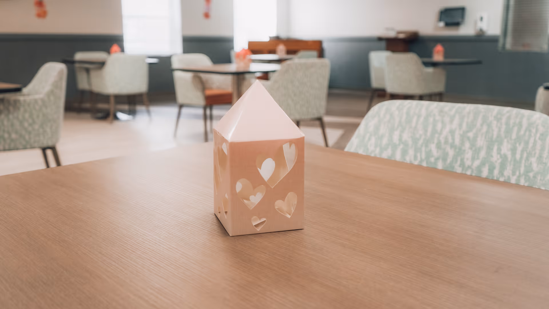 A wooden table with a small decorative house featuring heart-shaped cutouts in the center. In the background, there are several tables and cushioned chairs in a softly lit room with large windows and muted wall colors.