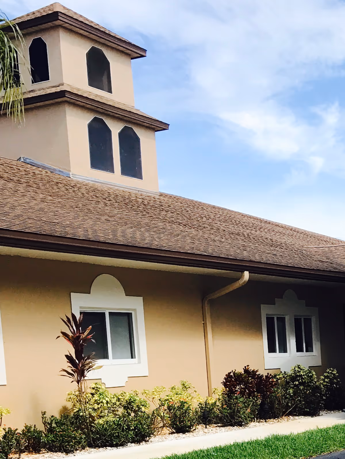 Beige stucco building exterior with a multi-tiered tower, shingled roof, windows, and landscaped shrubs.