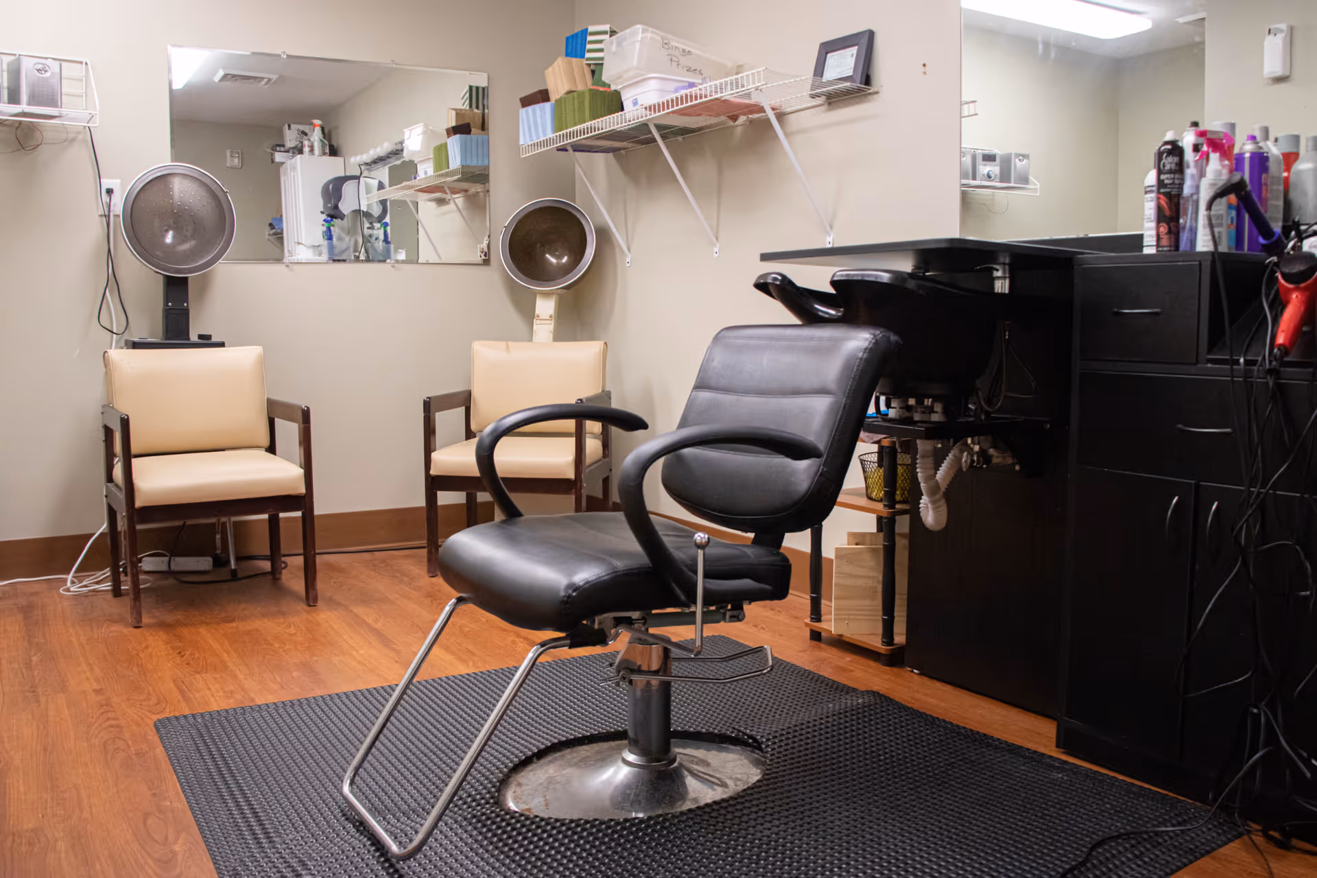 Interior view of a hair salon area in a senior living facility with a black salon chair in the center, two beige chairs with hair dryers behind them, a large mirror on the wall, and shelves holding hair care products and supplies.