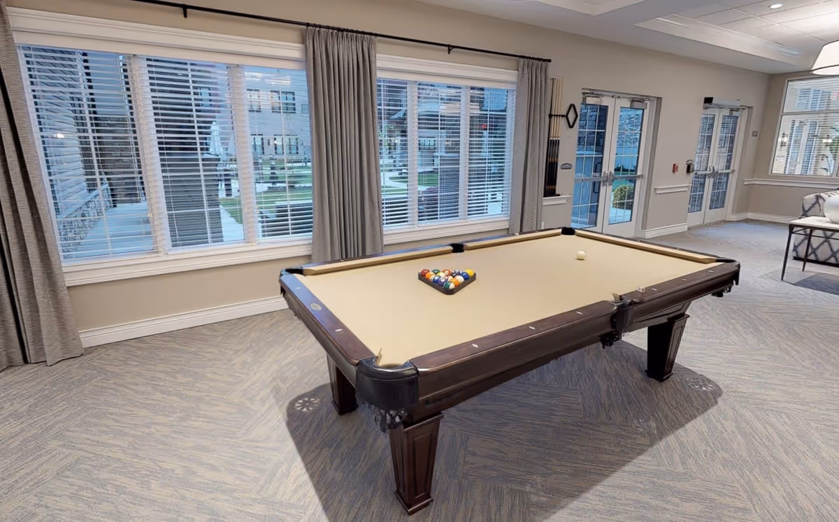 A spacious room with a beige pool table set up for a game, featuring a rack of billiard balls and a cue ball. The room has large windows with blinds and curtains, allowing natural light to enter. There are double glass doors leading outside, and a seating area with a sofa and a side table is partially visible on the right. The floor is carpeted with a subtle pattern, and the walls are painted in a neutral tone.