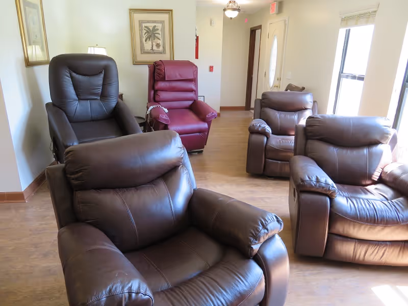 A bright room with five comfortable recliner chairs arranged on a wooden floor. The chairs are mostly dark brown leather, with one maroon recliner near the back. The room has light-colored walls, a framed picture, a lamp, and a door with windows letting in natural light.
