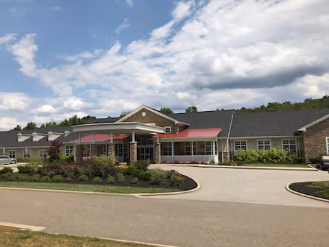 Front entrance of a single-story senior living facility with a covered portico, landscaped flower beds, and a circular driveway under a partly cloudy sky.