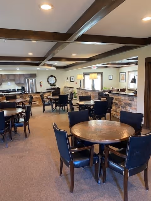Interior view of a communal dining area with multiple round wooden tables and black cushioned chairs. The room features exposed wooden ceiling beams, stone accent walls, and a kitchen area with stainless steel appliances in the background. The space is well-lit with recessed lighting and table lamps.