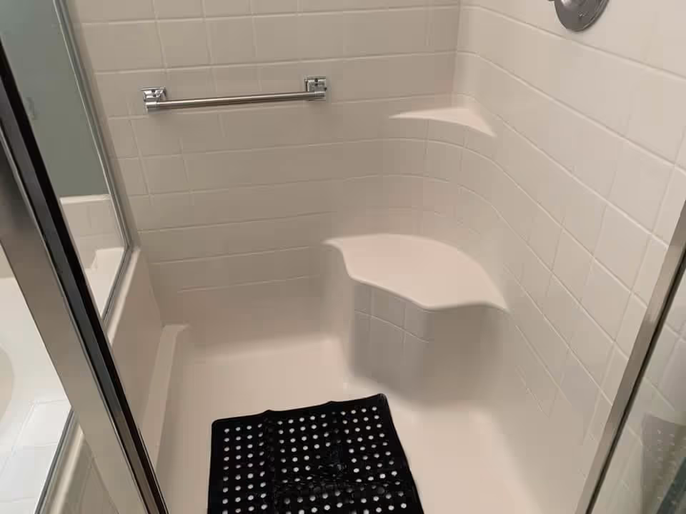 A white tiled shower area with a built-in corner seat and a metal grab bar on the wall. There is a black non-slip mat on the shower floor and a glass shower door partially visible on the left side.