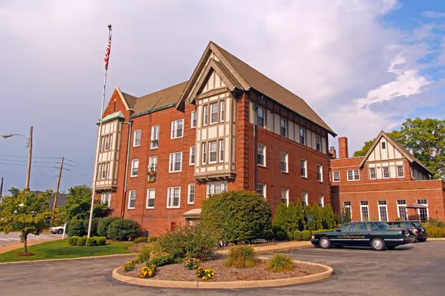 Exterior view of a large, multi-story brick building with Tudor-style architectural elements, surrounded by landscaping and a parking area with cars. An American flag is flying on a flagpole in front of the building under a partly cloudy sky.