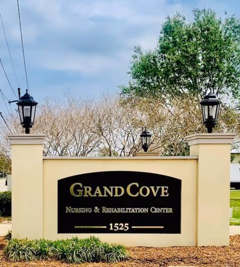Stone entrance sign reading "Grand Cove Nursing & Rehabilitation Center 1525" flanked by lamp posts and landscaping.