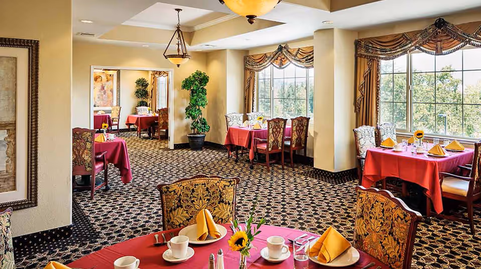 A dining room with several tables covered in red tablecloths, each set with yellow folded napkins, white cups, plates, and silverware. The room has large windows with decorative curtains letting in natural light, patterned carpet, and upholstered chairs with floral designs. There are potted plants and framed artwork on the walls.