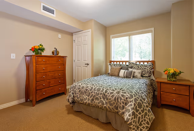 A cozy bedroom with a double bed covered in a patterned gray and white comforter and multiple pillows. There is a wooden dresser with flowers and a decorative item on top to the left, and a matching wooden nightstand with a vase of flowers to the right. A window with blinds is behind the bed, letting in natural light. The walls are painted beige and the floor is carpeted.