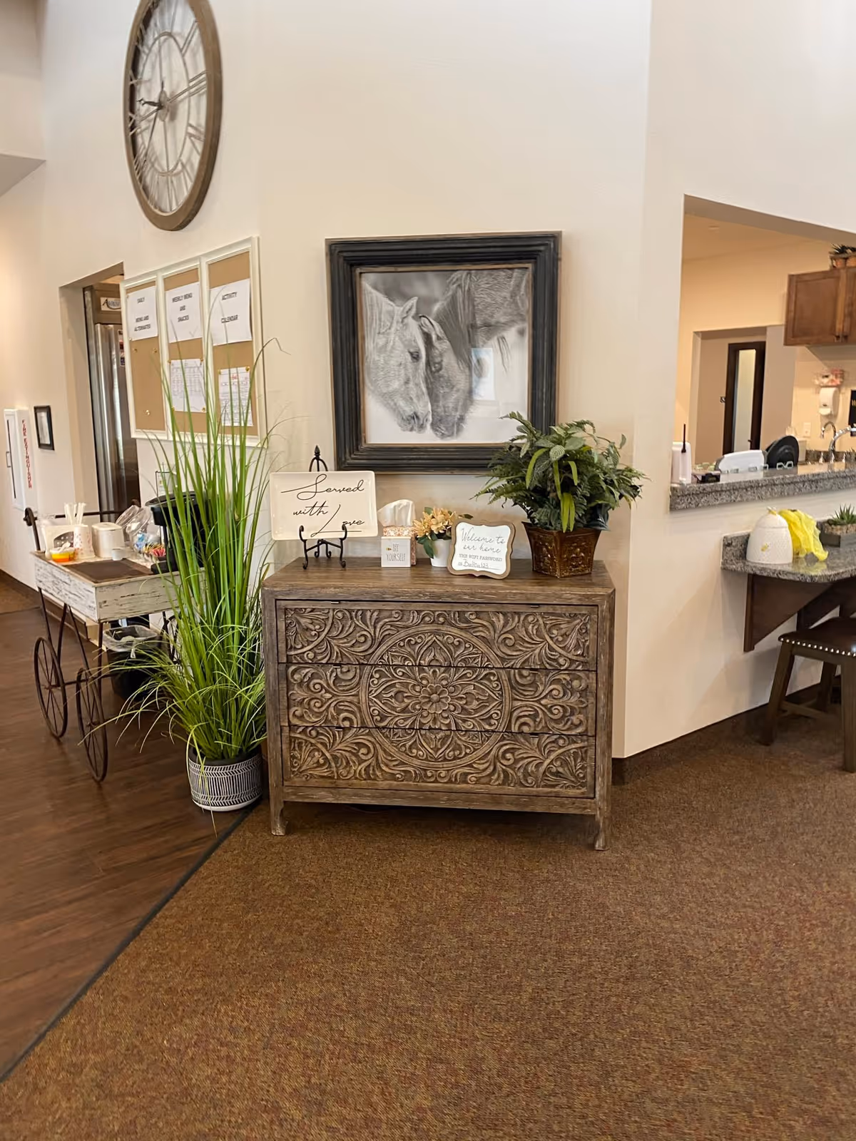 Interior view of a senior living facility showing a decorative wooden cabinet with intricate carvings, topped with potted plants and framed signs. Above the cabinet is a framed black and white picture of two horses touching heads. To the left, there is a tall green plant in a pot and a cart with various items. A large wall clock is visible on the wall, and to the right, a granite countertop with a chair is partially visible.