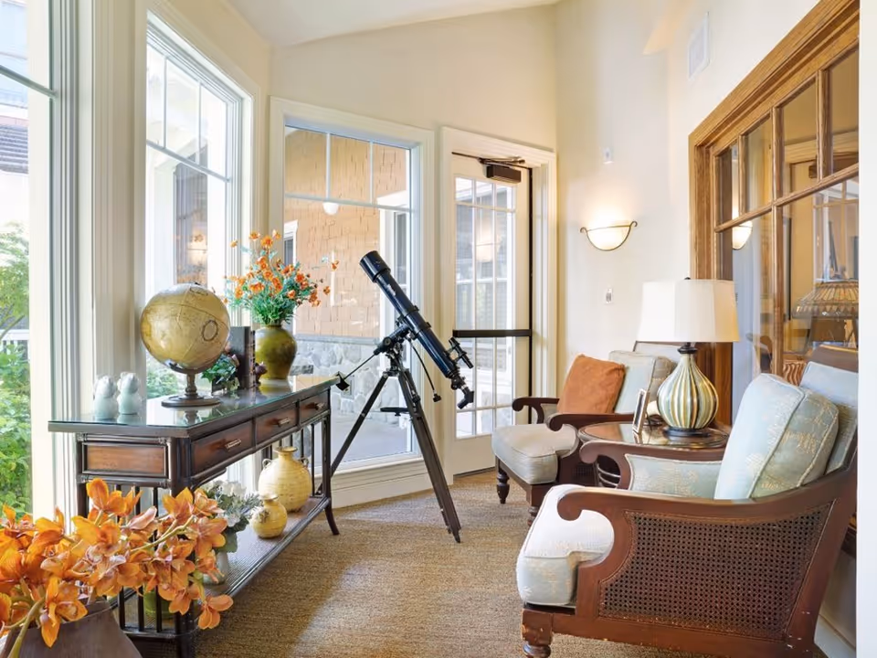 Bright sitting area with two upholstered armchairs, a telescope, and a console table with a globe and flowers by large windows.