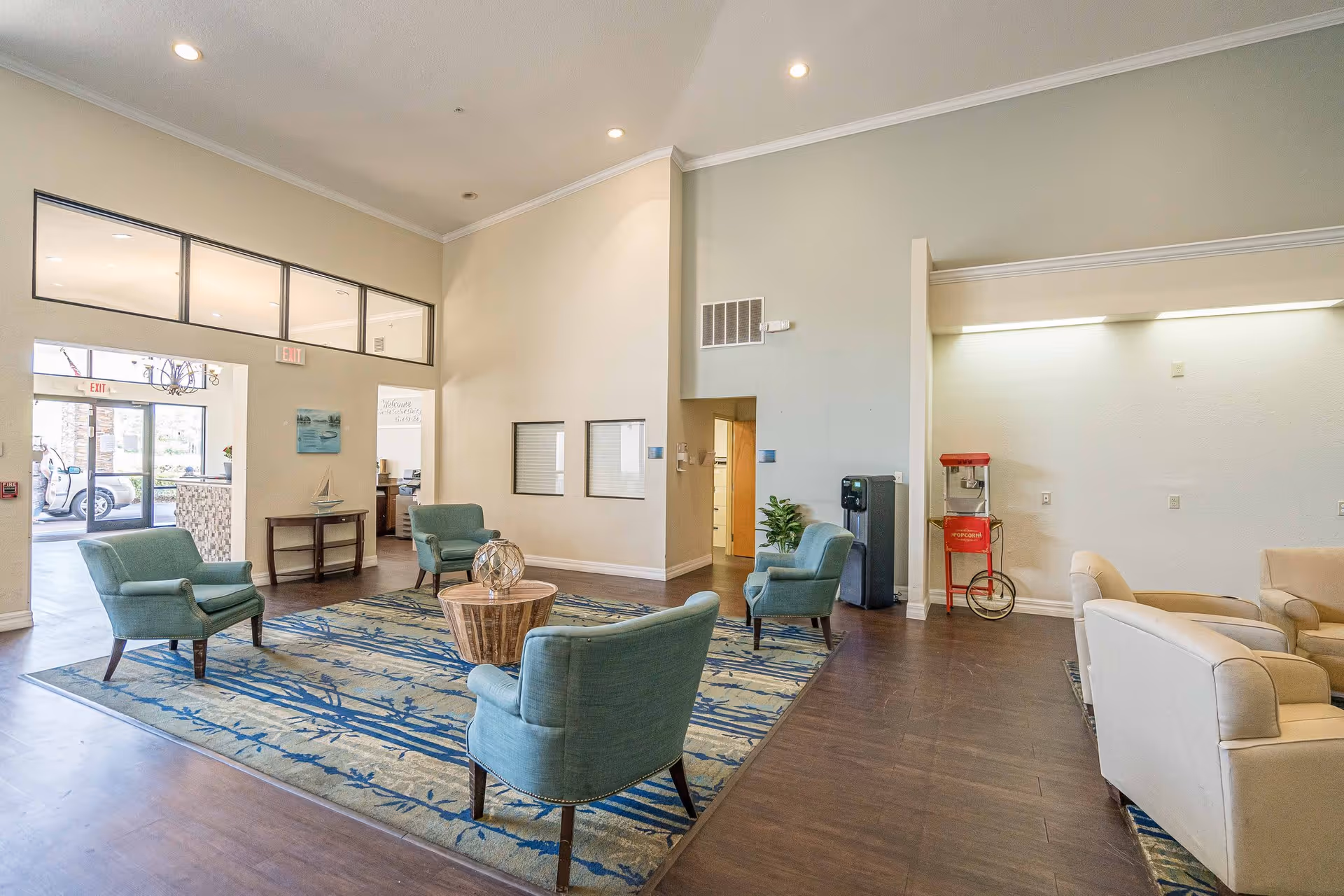 A spacious assisted living facility common area with four teal armchairs arranged around a round wooden coffee table on a patterned blue and beige rug. The room has high ceilings, light-colored walls, and wood flooring. There is a popcorn machine and a water dispenser against one wall, and a beige sofa set is partially visible on the right side. Large windows and glass doors provide natural light and a view of the outside.