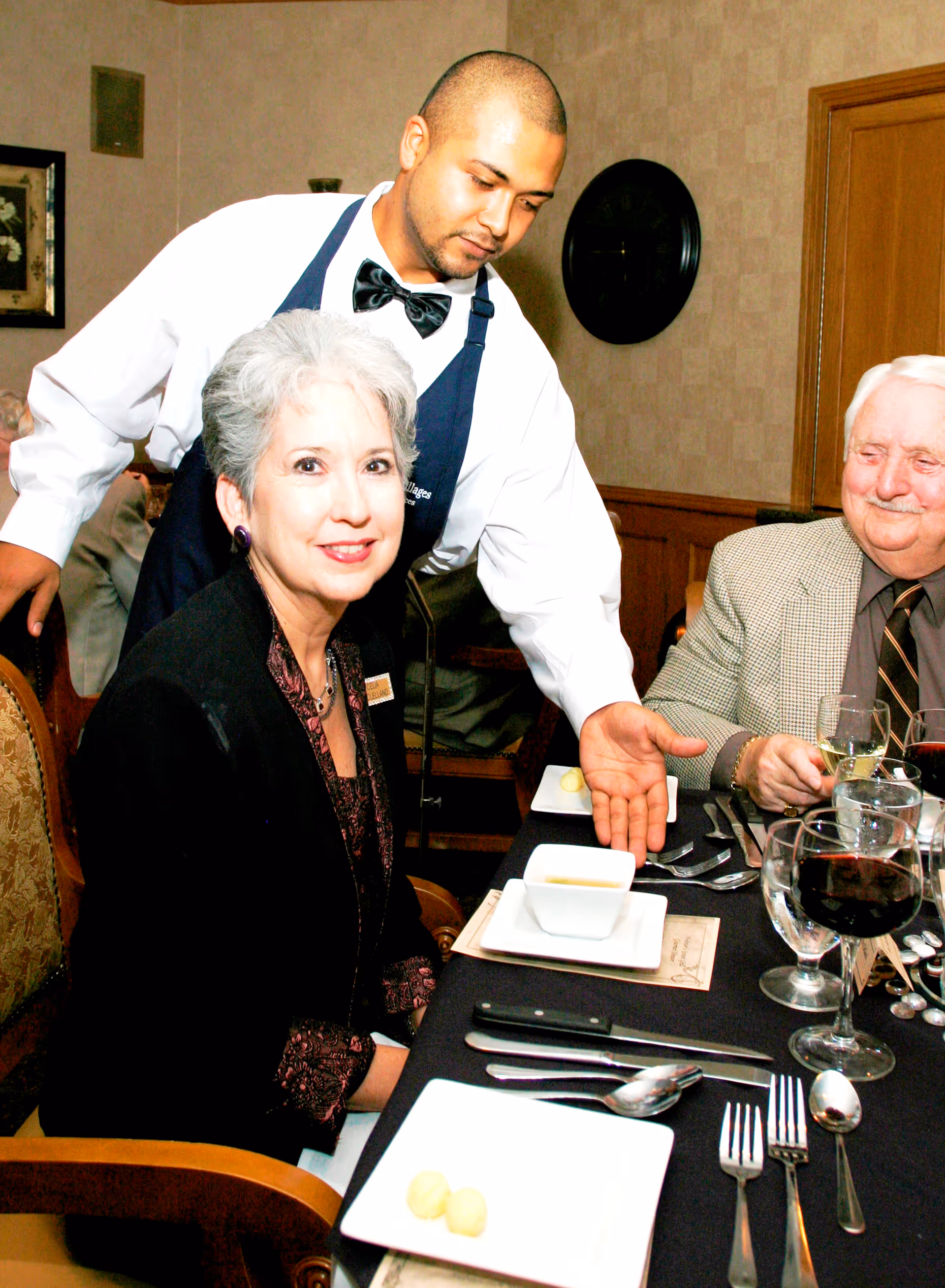 A waiter dressed in a white shirt, black bow tie, and dark apron is serving a bowl of soup to a seated elderly woman with short gray hair wearing a black jacket. An elderly man with white hair and a mustache, dressed in a checkered blazer and dark tie, is seated next to her at a dining table set with glasses of wine, water, and silverware.