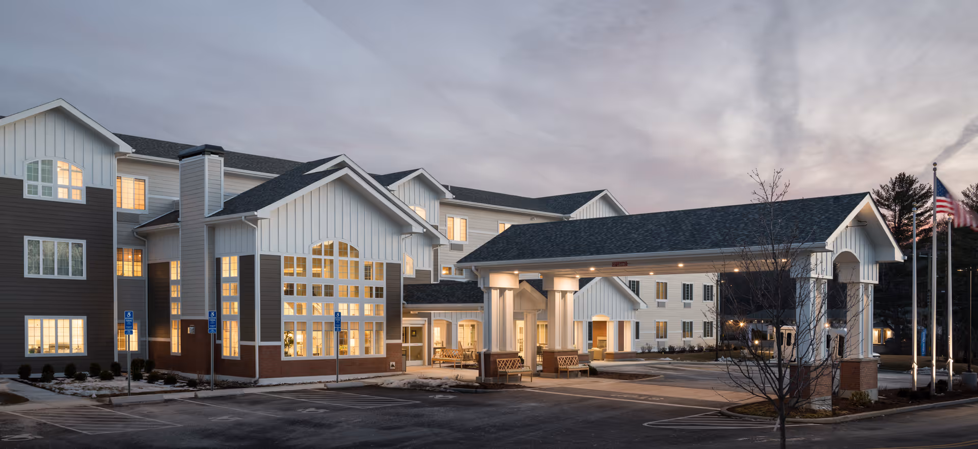Exterior view of a senior living facility building at dusk with lights on inside. The building has multiple stories with large windows and a covered entrance with columns. There are parking spaces and an American flag on a flagpole near the entrance.