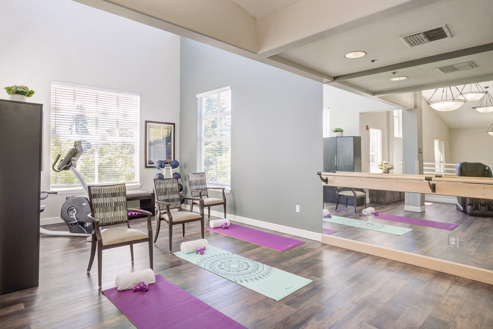 A bright exercise room with large windows, two yoga mats on the floor each with a towel and small dumbbells, two chairs, an exercise bike, a set of dumbbells on a rack, and a large wall mirror with a ballet barre.