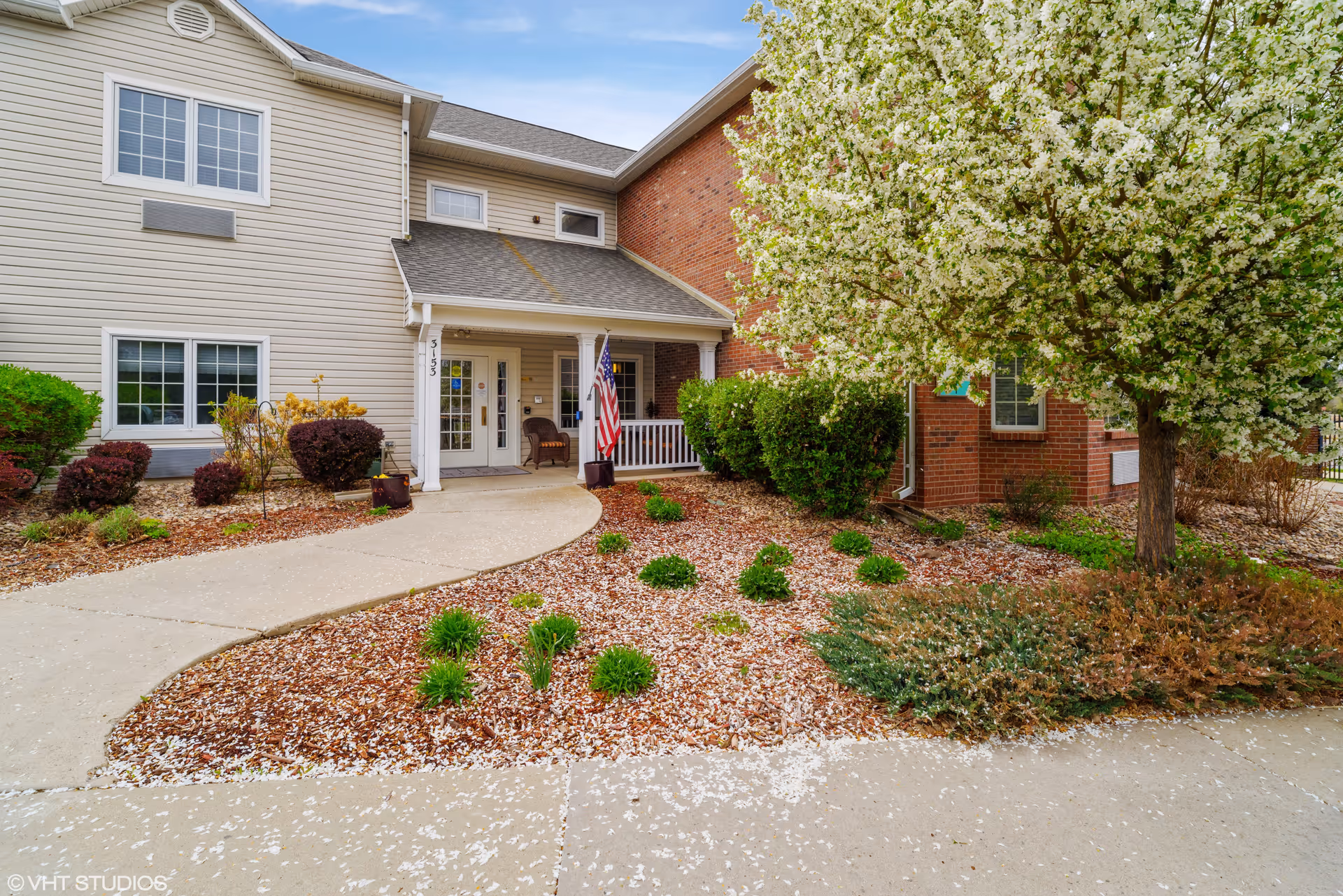 Entrance to a senior living facility with a covered porch, American flag, and landscaped garden featuring bushes, small plants, and a tree with white blossoms. The building has beige siding and red brick exterior walls.