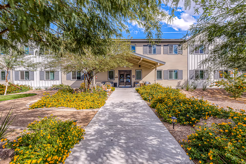 Front exterior view of a two-story senior living facility building with beige and white siding, multiple windows, and a covered entrance. The walkway leading to the entrance is flanked by landscaped gardens with yellow flowers and trees providing shade. The sky is blue with some clouds.