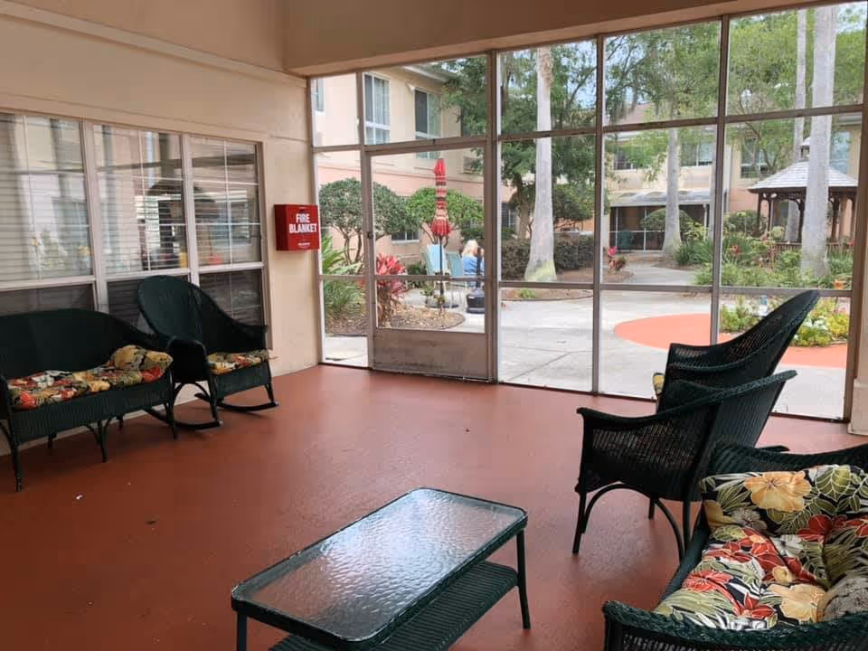 A screened-in porch area with green wicker chairs and a glass-top coffee table. The chairs have floral cushions. Outside the porch, there is a courtyard with trees, plants, a gazebo, and a person sitting on a bench.