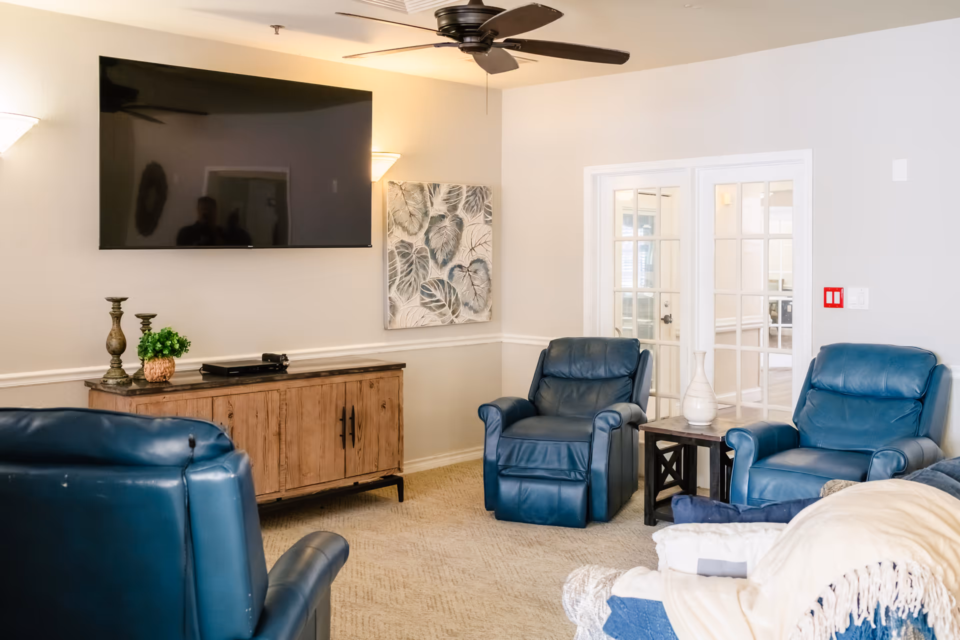 A cozy common room with blue recliner chairs, a wall-mounted TV, a wooden media cabinet, and a ceiling fan.