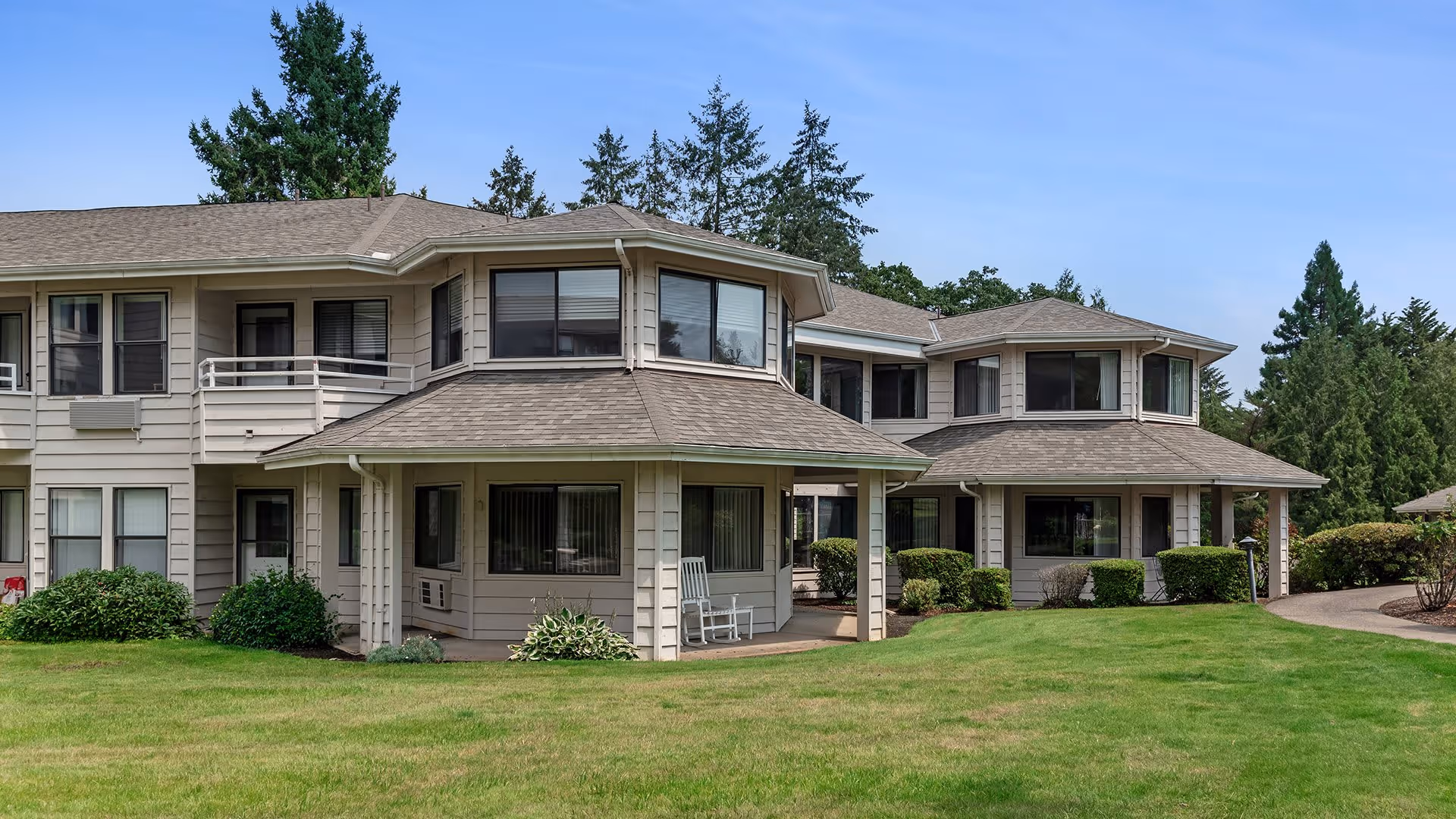 Exterior view of a two-story senior living facility building with beige siding and multiple windows. The building is surrounded by a well-maintained green lawn, bushes, and tall trees under a clear blue sky. There is a white rocking chair on a covered porch area.