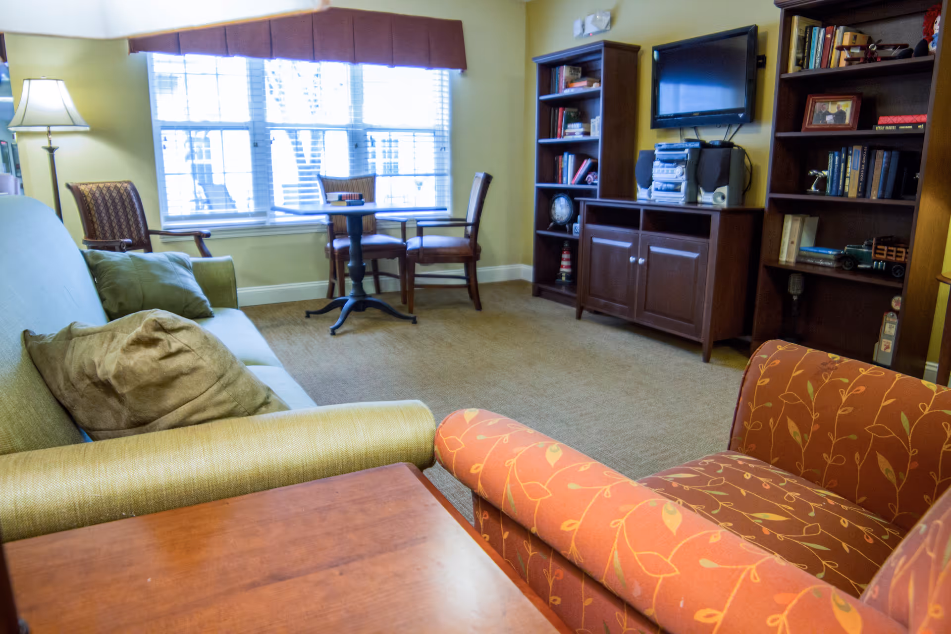 A cozy living room with a green sofa, an orange armchair with leaf patterns, a wooden coffee table, and a floor lamp. There is a window with blinds and a valance, two wooden chairs with a small round table between them, and a wooden entertainment center with shelves holding books, a stereo system, and a wall-mounted TV.