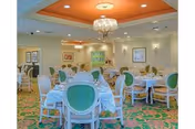 A spacious dining room with multiple round tables covered with white tablecloths and set with plates and silverware. The chairs have white frames and green cushioned backs and seats. The room features a patterned carpet with green and orange colors, a chandelier hanging from an orange ceiling section, and framed artwork on the walls.