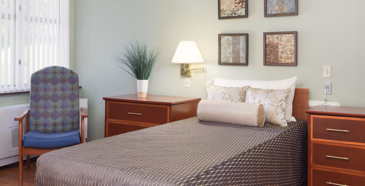 A tidy, well-lit bedroom with a single bed covered in patterned bedding, two wooden nightstands, a wall lamp, framed artwork, and an upholstered chair by the window.