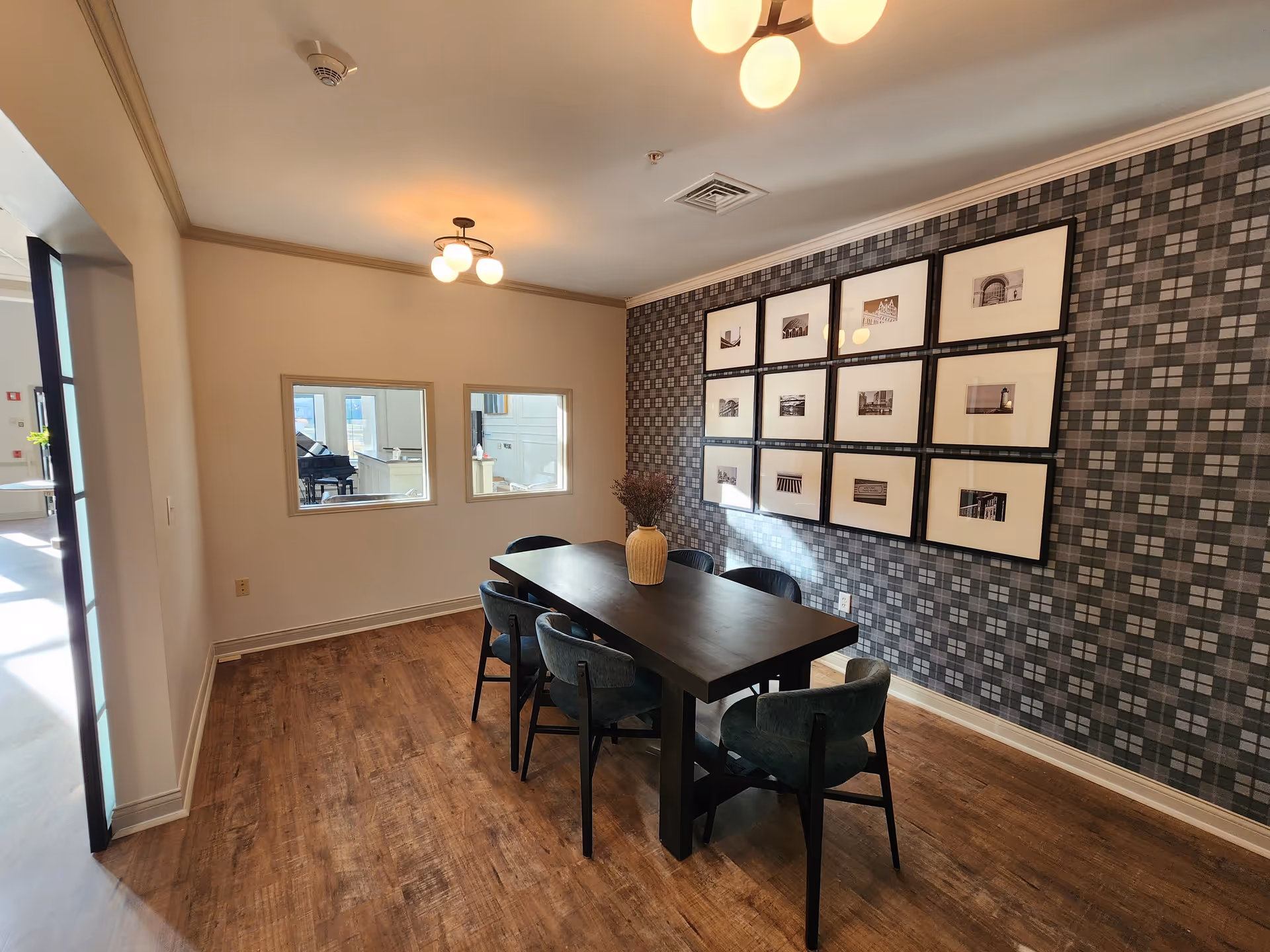 A small dining area with a dark wooden table and six upholstered chairs. A vase with dried flowers is placed on the table. The wall on the right side features a patterned wallpaper with a grid of twelve framed black and white photographs. Two small windows on the far wall look into another room with a piano visible. The floor is wooden, and the ceiling has two modern light fixtures.