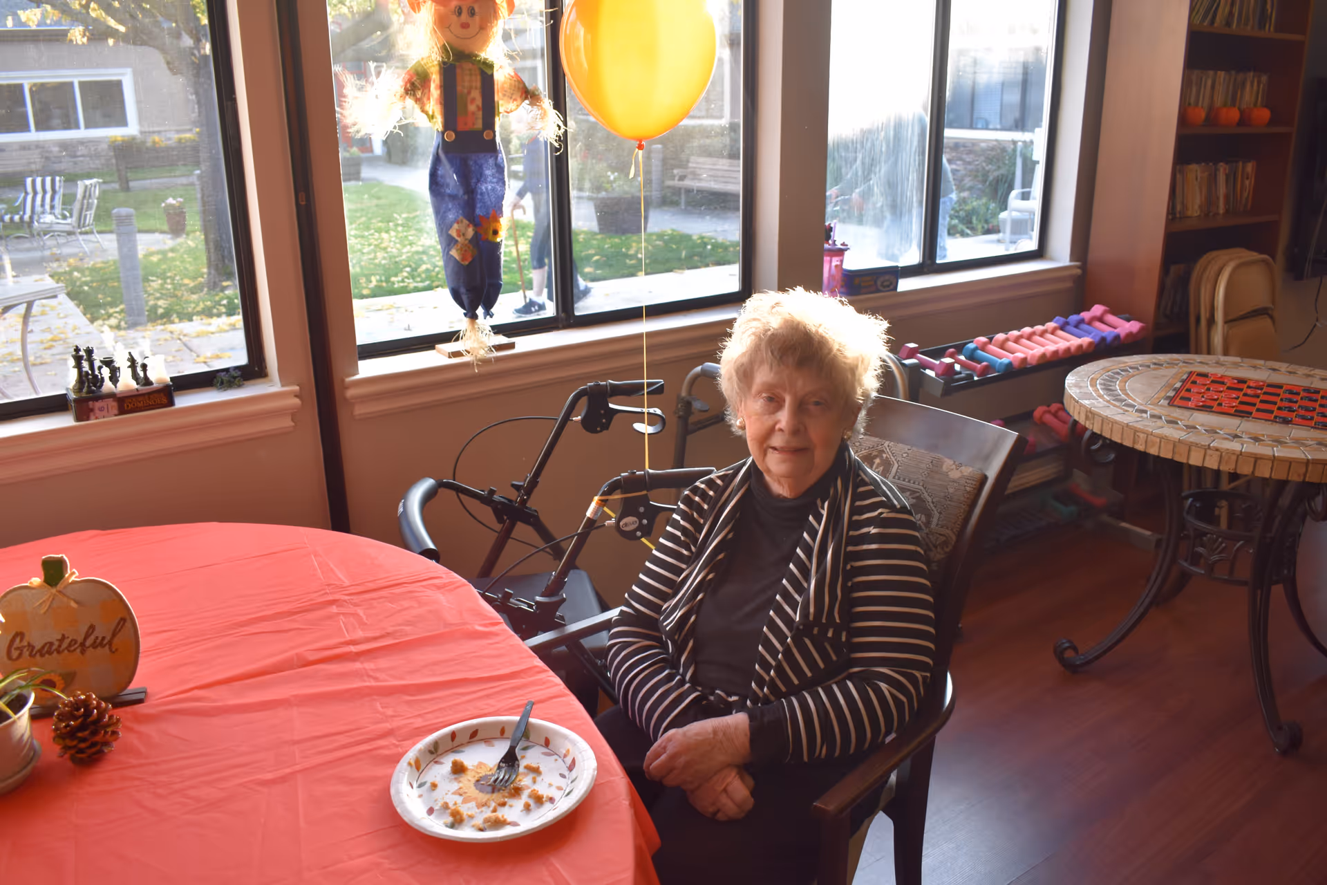 An elderly woman sitting at a table with a red tablecloth in a well-lit room. There is a partially eaten piece of cake on a paper plate in front of her. Behind her is a walker, a yellow balloon, a scarecrow decoration hanging in the window, and a rack of colorful dumbbells. The room has large windows showing an outdoor patio area with chairs and greenery.